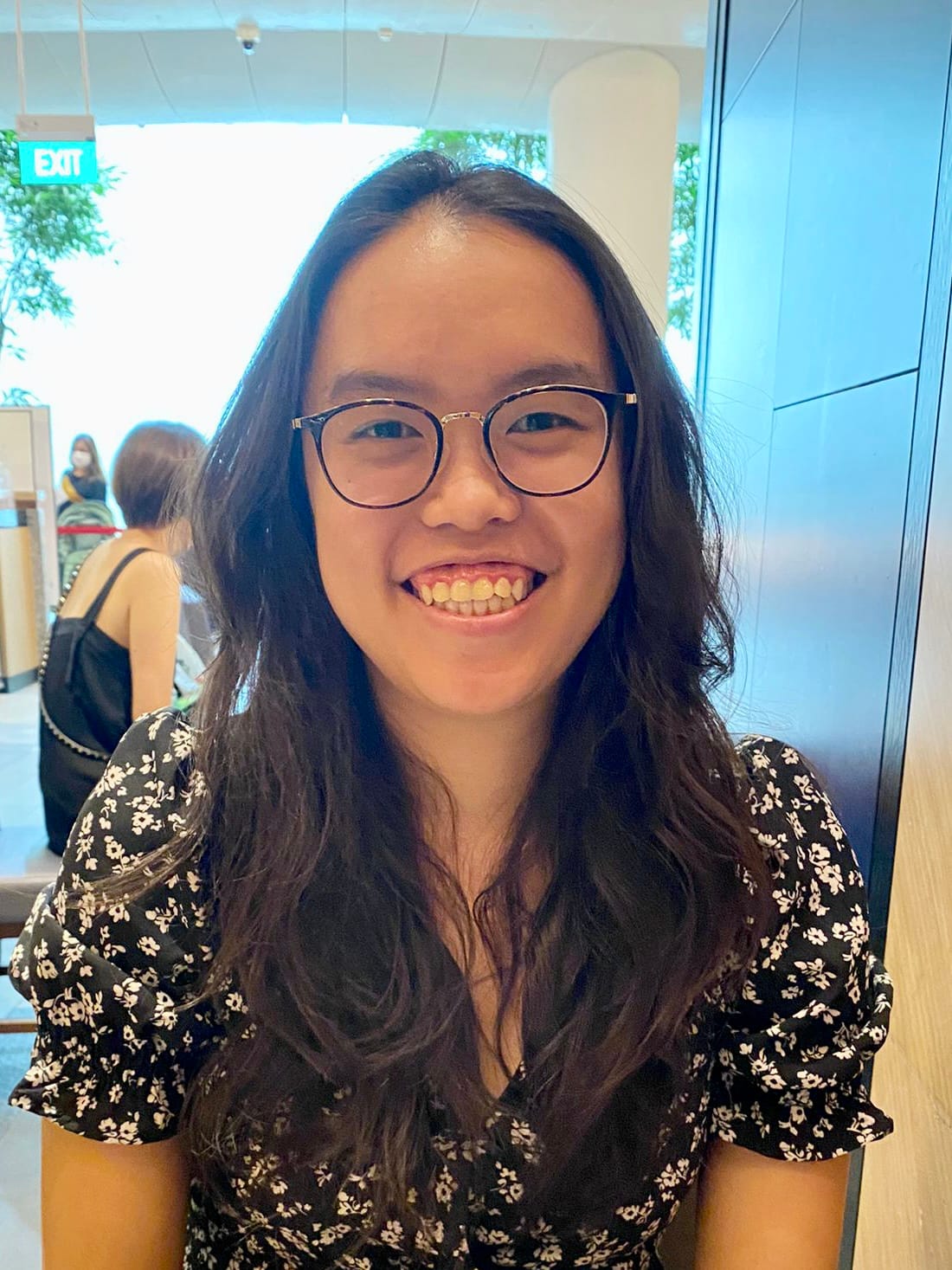 A young woman with long dark hair and round glasses smiles broadly, wearing a black floral dress indoors.
